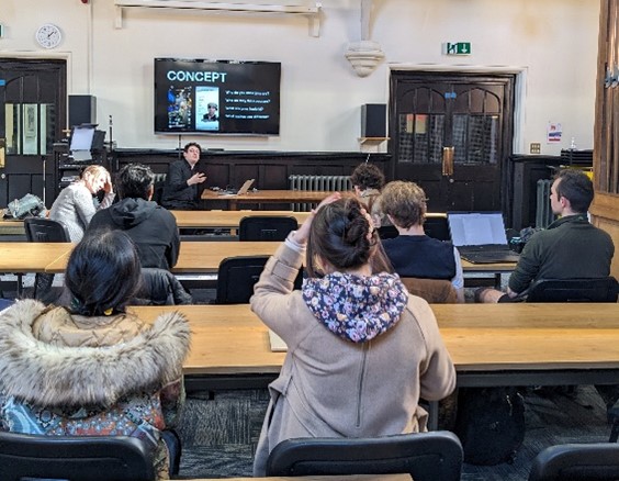 Students sitting in a lecture room at tables watching a presentation being given by a lecturer